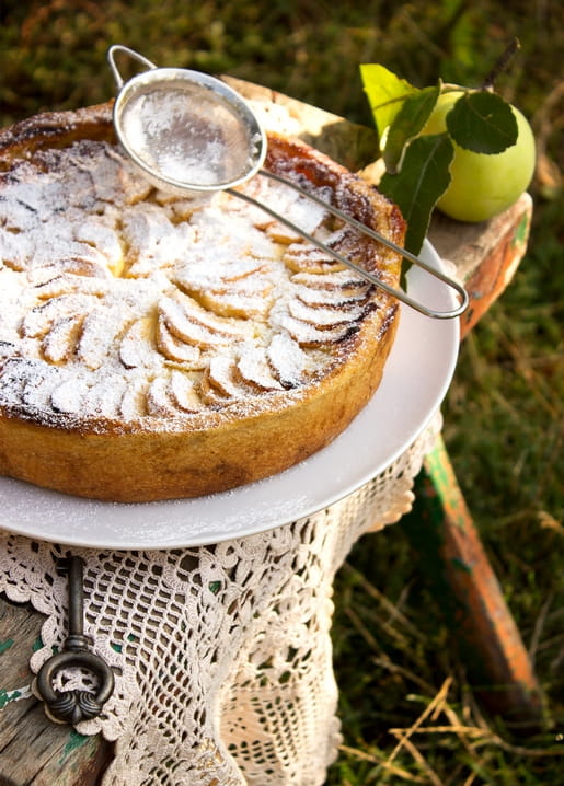 Apple tart dusted with powdered sugar and a fresh apple on a lace-covered garden stool