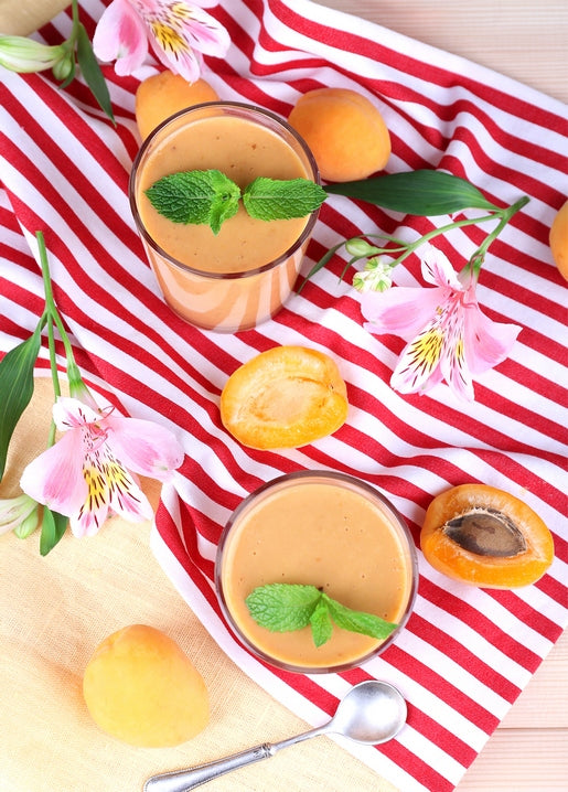 Apricot smoothie with mint garnish, ripe apricots, and pink flowers on red-striped cloth