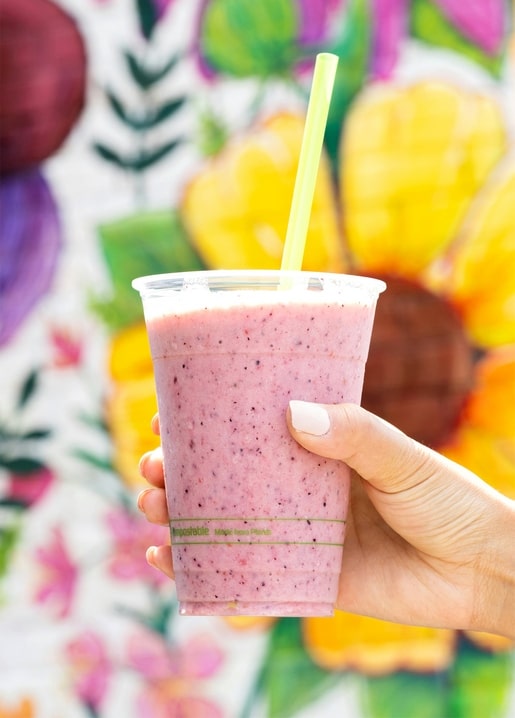 Hand hält einen pinken Beeren-Smoothie mit grünem Strohhalm vor buntem Blumen-Wandbild