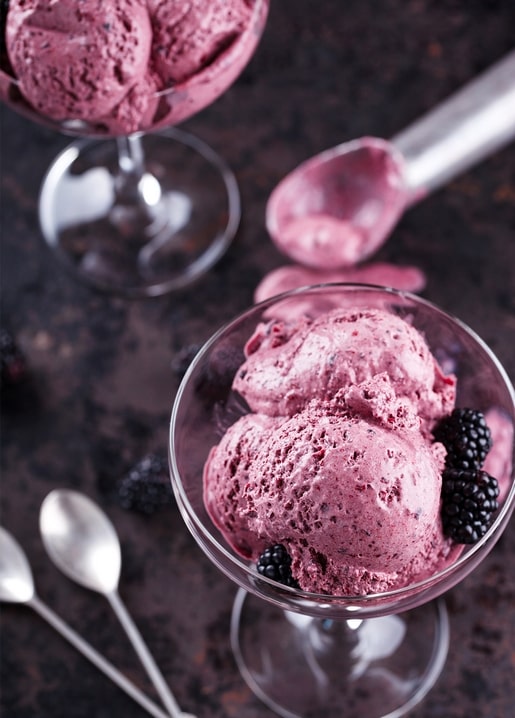 Brombeereis in Cocktailgläsern mit frischen Beeren und Silberlöffeln auf dunklem Stein von oben