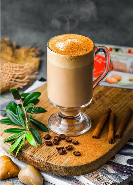 Dampfender Zimt-Latte im Glas auf Holzbrett mit Kaffeebohnen und Zimtstangen