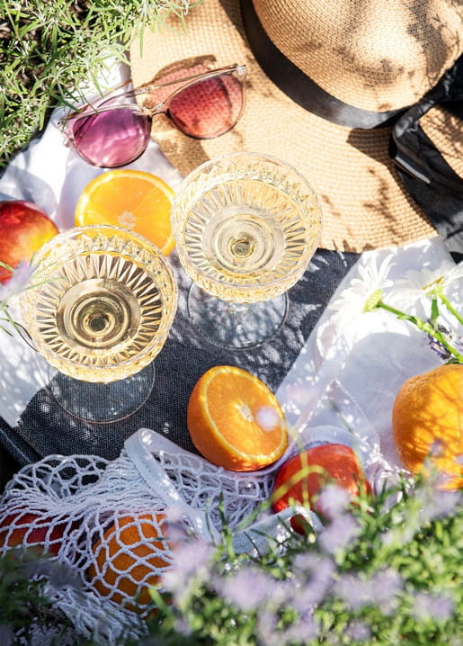 Sektgläser, Orangenscheiben und ein Strohhut auf einer Leinen-Picknickdecke