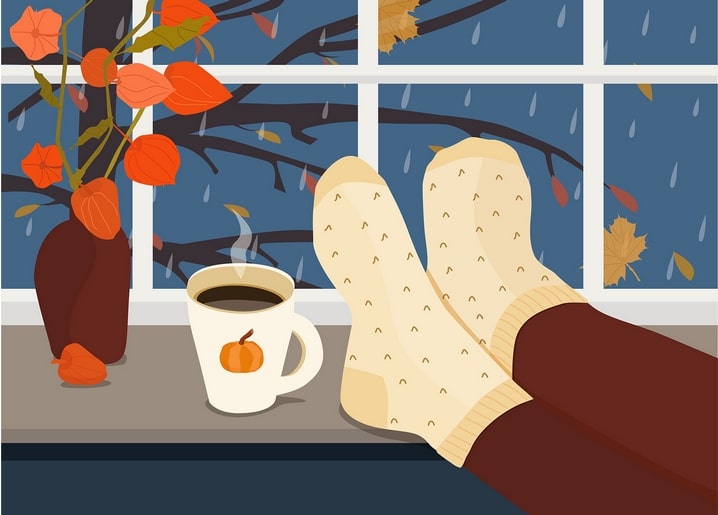 Feet in wool socks on a windowsill beside a pumpkin mug and orange physalis in the rain