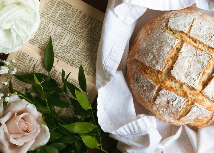 Frischer Brotlaib auf weißem Leinen neben Rosen und einem aufgeschlagenen alten Buch