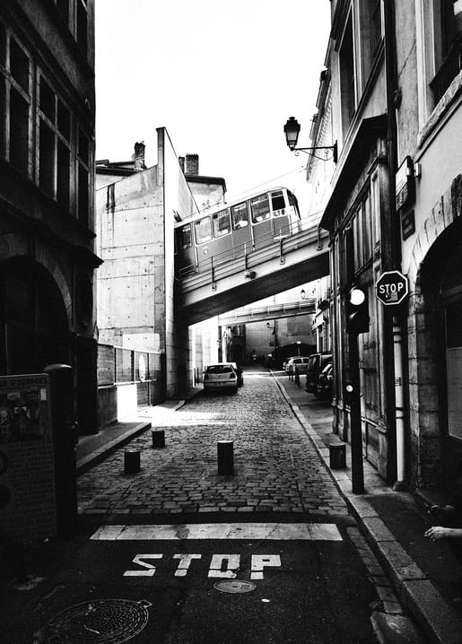 Schwarz-weißes Foto einer Hochbahn über einer schmalen Kopfsteinpflastergasse mit STOP-Schriftzug auf der Straße.