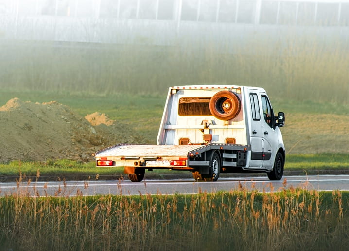 White flatbed truck hauling a small wooden cabin down a misty country road at dawn
