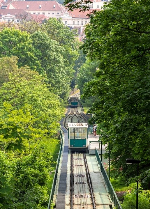Green funicular railway cars climbing through a forest toward a hilltop town with red rooftops