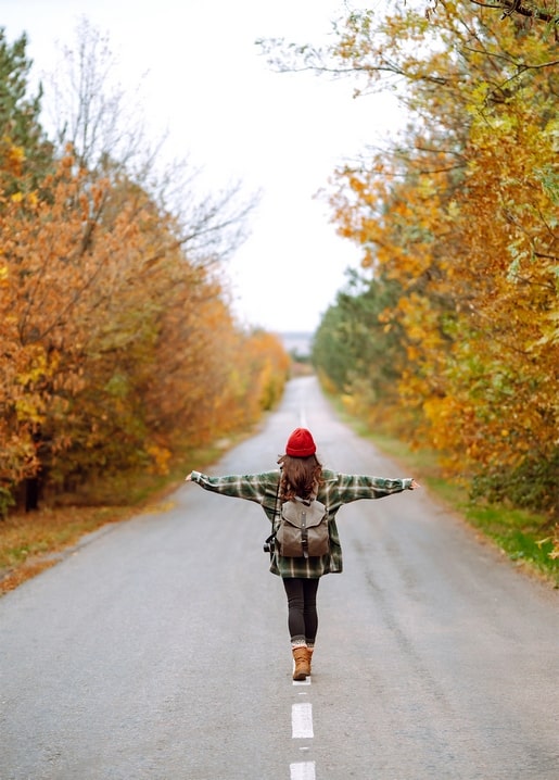 Mädchen mit roter Mütze und Rucksack geht mit ausgebreiteten Armen eine Herbststraße entlang