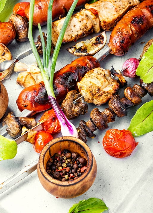 Gegrillte Spieße mit Würstchen, Hähnchen, Pilzen und Tomaten neben einer Schale Pfefferkörner