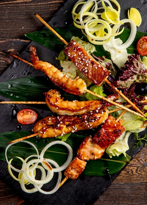 Grilled shrimp, chicken and bacon skewers with onion rings and greens on a bamboo leaf