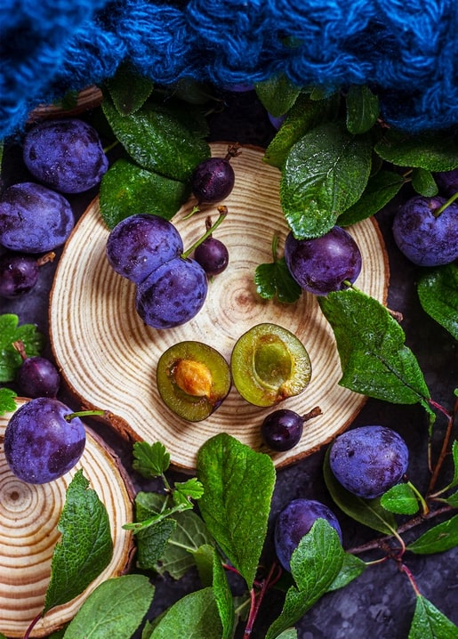 Halved purple plums and whole fruit on a wood slice with green leaves