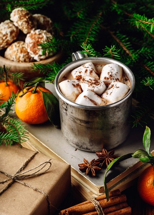 Tin mug of cocoa topped with marshmallows next to tangerines and gingerbread cookies