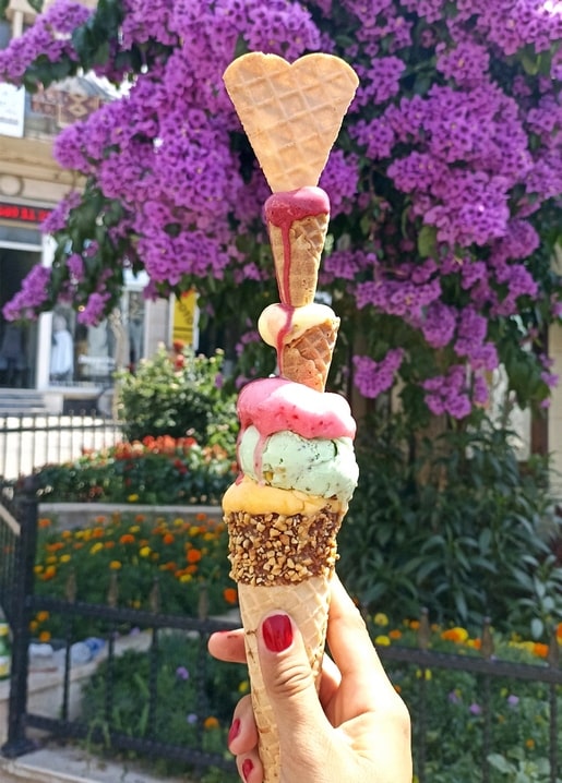 Hand hält eine mehrstöckige Eistüte mit Waffelherz vor violetter Bougainvillea
