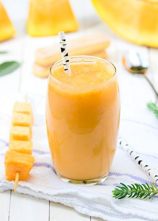 Glas Mango-Smoothie mit gestreiftem Strohhalm und Obstspieß auf gestreiftem Tuch
