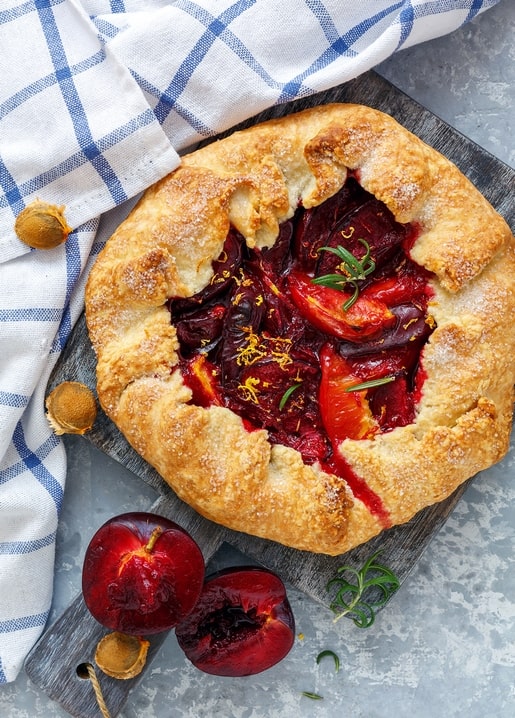 Baked plum galette topped with orange zest and rosemary on a blue checkered cloth