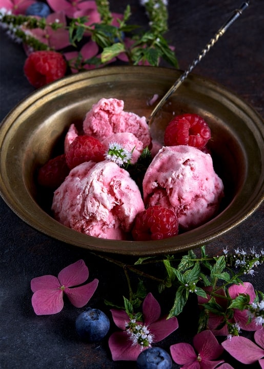 Himbeereis in Messingschale mit Himbeeren, Blaubeeren und rosa Blütenblättern