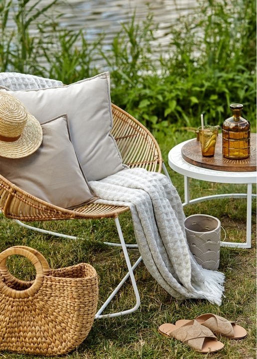 Rattan armchair with pillows and a straw hat beside a lake, wicker basket and drink on a small table