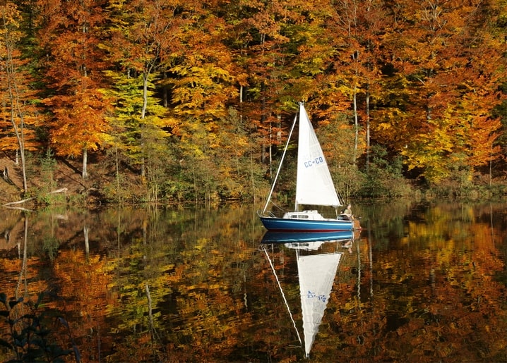 A sailboat on still water perfectly mirrored beneath a shoreline of red and orange autumn trees