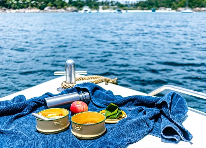 Zwei Lunchboxen, ein Apfel und eine Thermoskanne am Bug eines Segelboots, Küste in der Ferne
