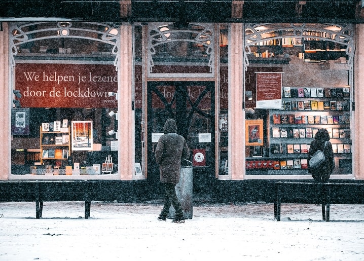 Snowy street with a lit bookshop window and two people walking in a snowstorm