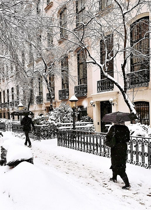 Two people walking under umbrellas on a snowy street lined with townhouses
