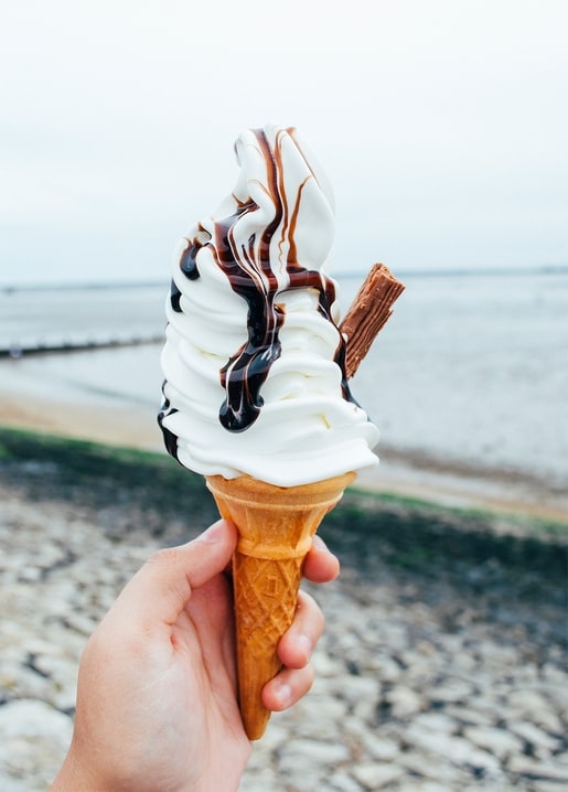 Hand hält Softeis-Waffel mit Schokoladensauce vor einem steinigen Strand