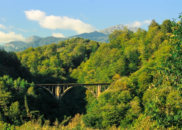 Stone arch bridge crossing a green valley surrounded by forested mountains