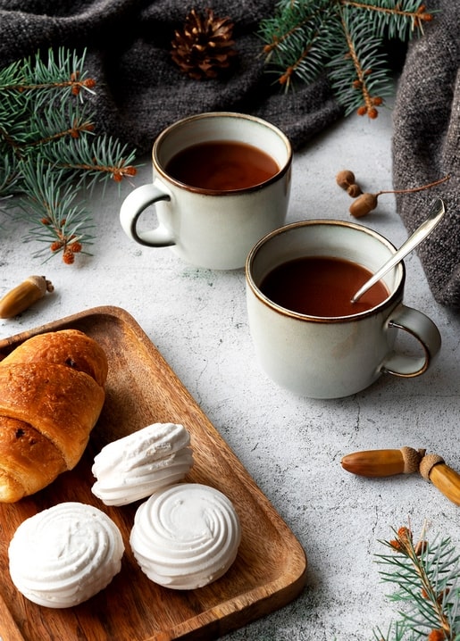 Two cups of tea with a croissant and meringue cookies among pine branches and a pinecone
