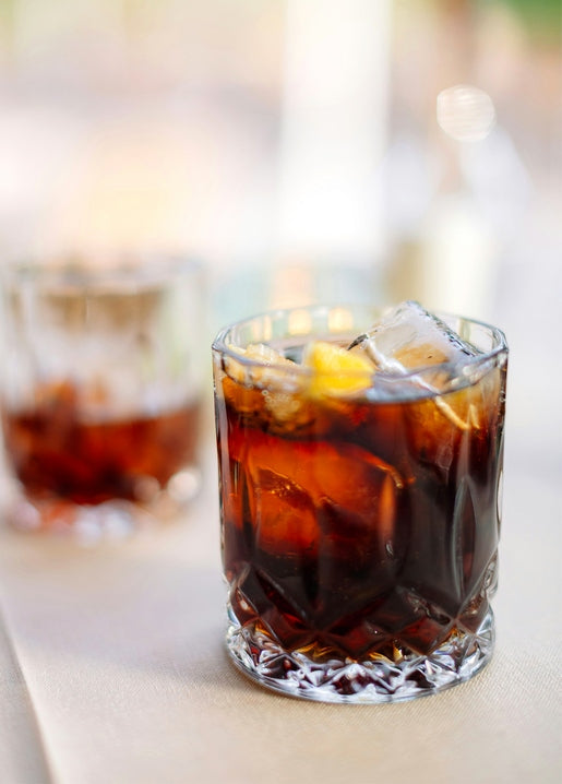 Glass of whiskey cola with ice cubes and a lemon slice, a second glass blurred in the background