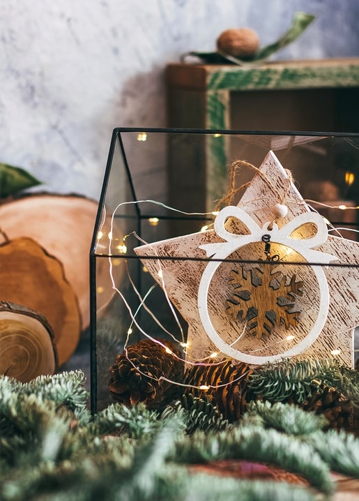 Wooden star ornament with snowflake cutout glowing inside a glass lantern with fairy lights and pinecones