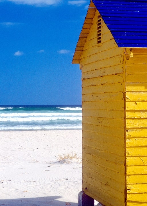 Weathered yellow beach hut with a blue roof facing white sand and turquoise sea waves