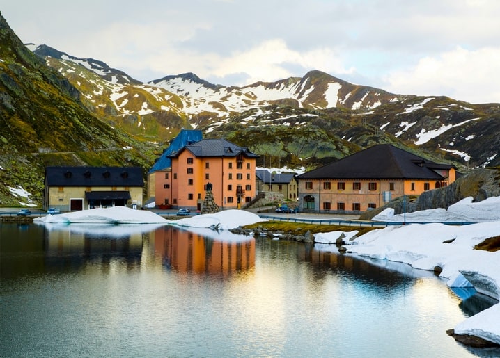 Ockerfarbene Alpengebäude mit dunklen Dächern spiegeln sich in einem See vor schneebedeckten Gipfeln.