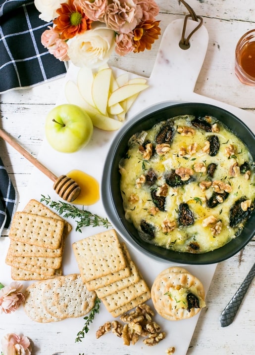 Baked cheese with walnuts and figs on a marble board, surrounded by crackers, apple slices, and flowers