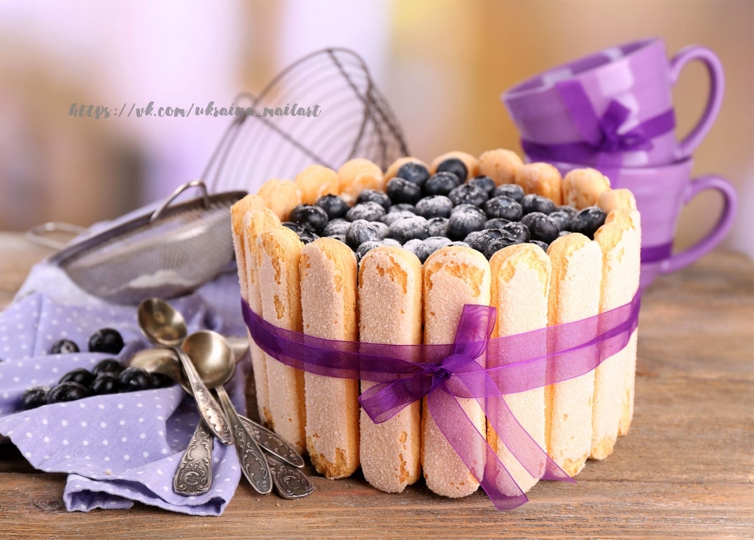 Charlotte-Torte mit Heidelbeeren und lila Schleife neben lila Teetassen