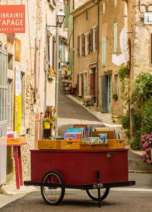 Червоний дерев'яний візок з книгами на бруківці біля невеликої книгарні