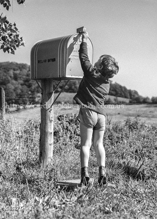 Black-and-white photo of a boy reaching up to mail a letter at a rural mailbox