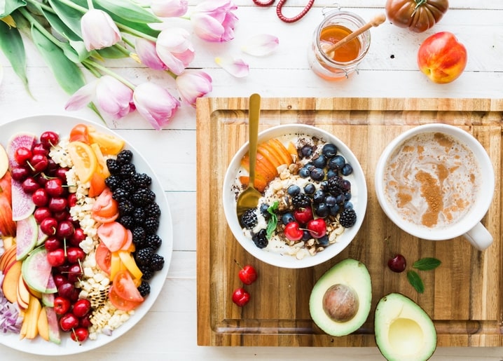 Smoothie bowl, fresh fruit, avocado, and pink tulips arranged on a wooden breakfast table