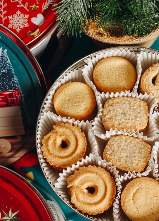 Offene Dose mit Butterkeksen neben weihnachtlichen Dosen und einem kleinen Tannenbaum