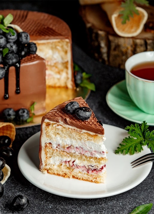 Cake slice with blueberries and chocolate glaze beside a cup of tea
