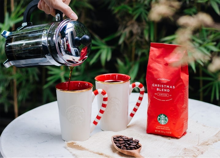 French Press gießt Kaffee in Tassen mit Zuckerstangen-Henkeln neben roter Kaffeetüte.