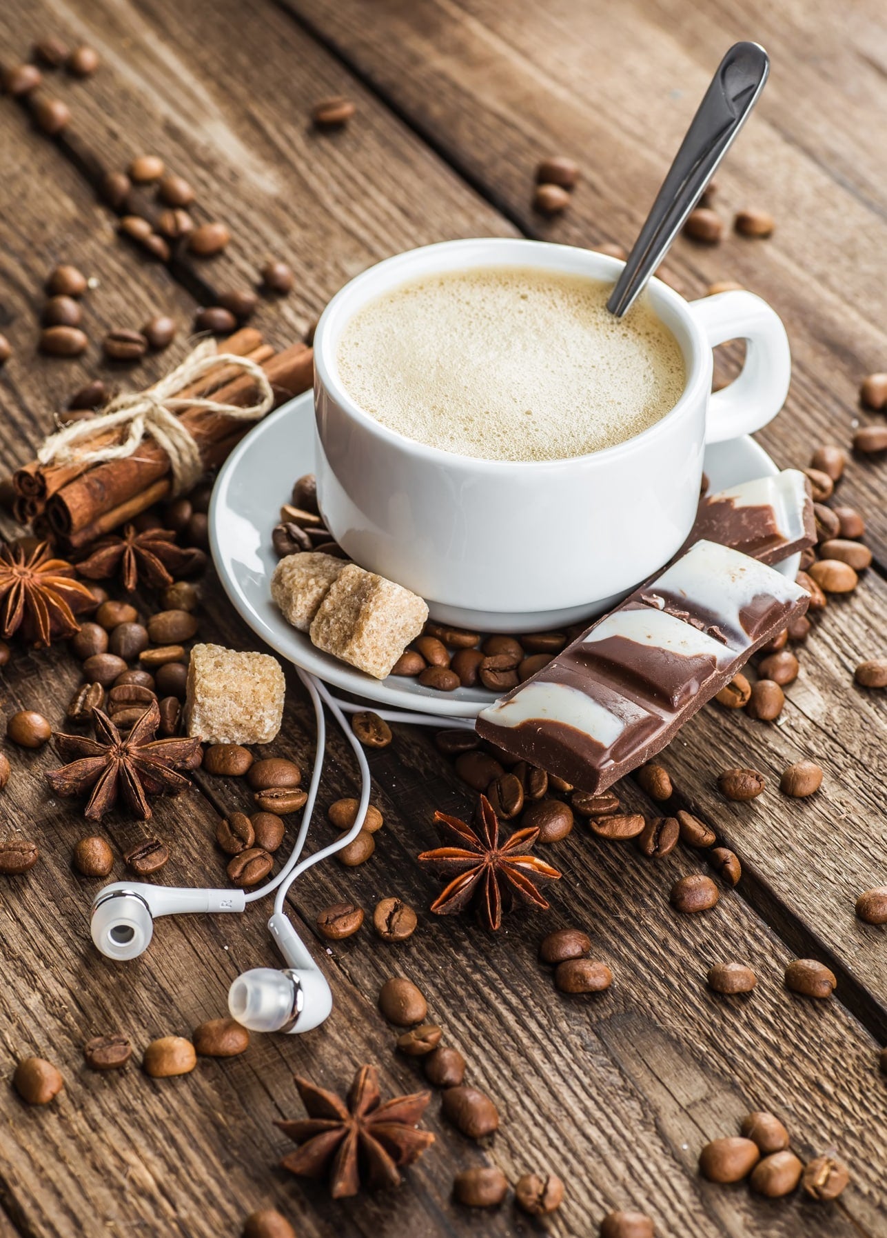 Cappuccino surrounded by coffee beans, cinnamon, star anise, chocolate and white earbuds