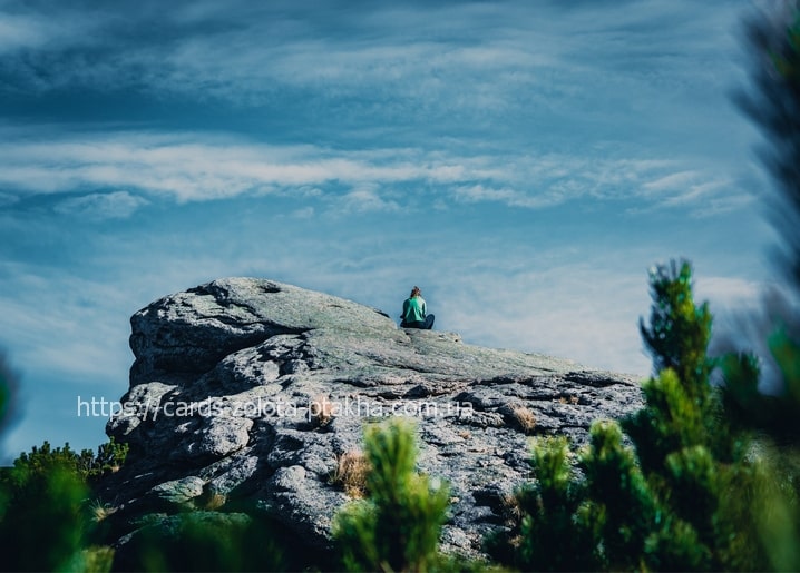 Hiker sitting cross-legged on a rocky Carpathian mountain peak under a blue sky with clouds