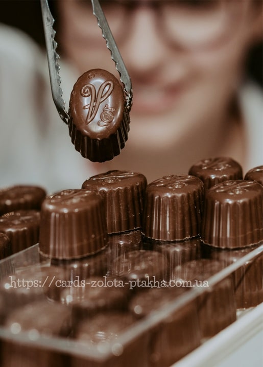 Zange hält eine Praline mit Monogramm über einer Schachtel glänzender Pralinen