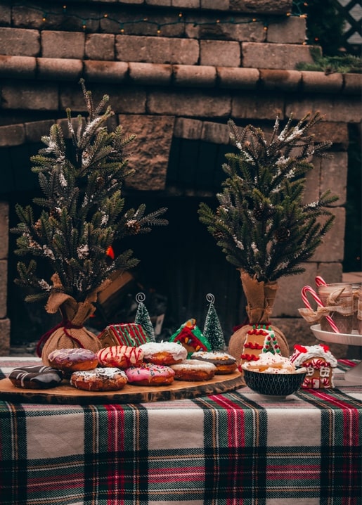 Frosted donuts on a wood board beside two snow-dusted mini pine trees and a stone fireplace with plaid tablecloth.