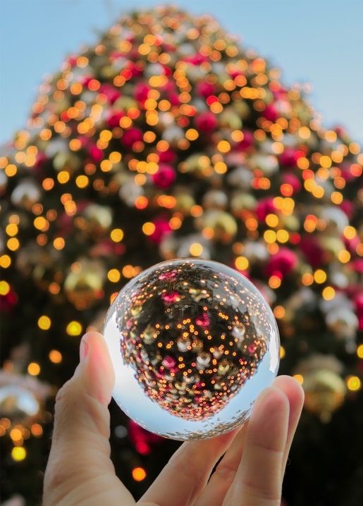 A hand holding a glass sphere reflecting a lit Christmas tree against a blue sky