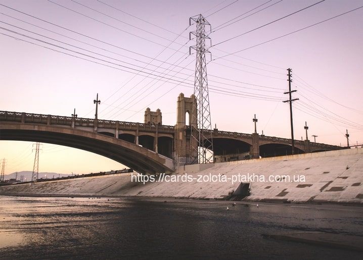 Blassgraue Betonbogenbrücke über einen Flusskanal in der Dämmerung mit hohem Strommast im Hintergrund.