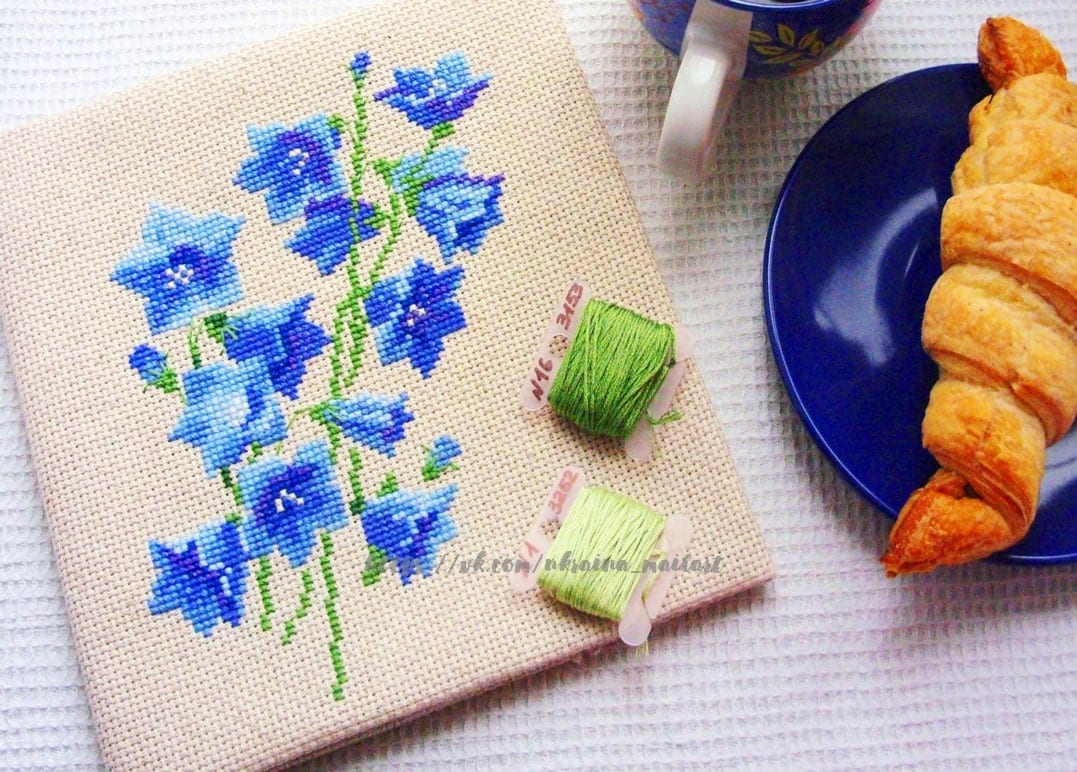 Kreuzgestickte blaue Glockenblumen neben Croissant und einer Tasse Kaffee