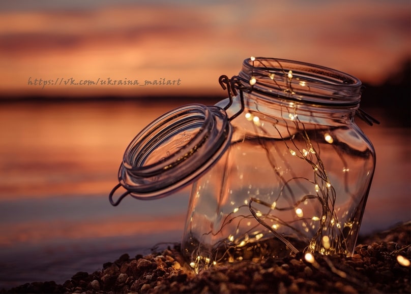 Einmachglas mit warmer Lichterkette auf Kieselsteinen am Wasser bei Sonnenuntergang