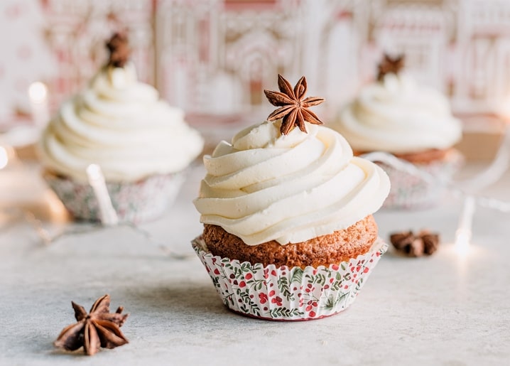 Cupcake with whipped frosting and star anise, Christmas-patterned wrappers and lights blurred behind