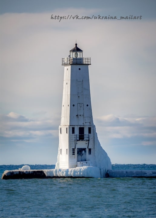Weißer Leuchtturm mit vereistem Sockel unter grauem Winterhimmel